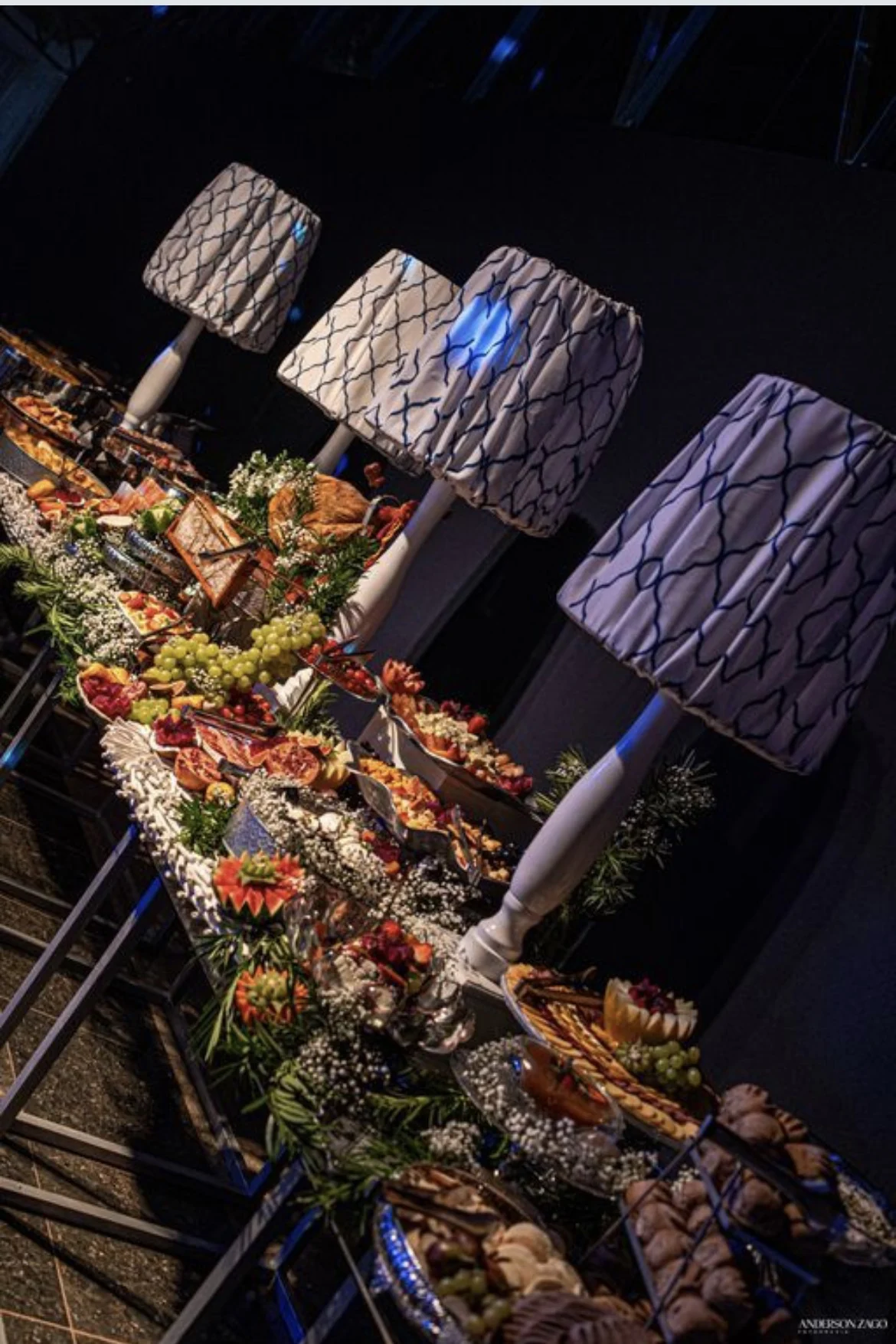 Mesa longa de buffet de casamento com frutas, travessas e luminárias acesas ao fundo.