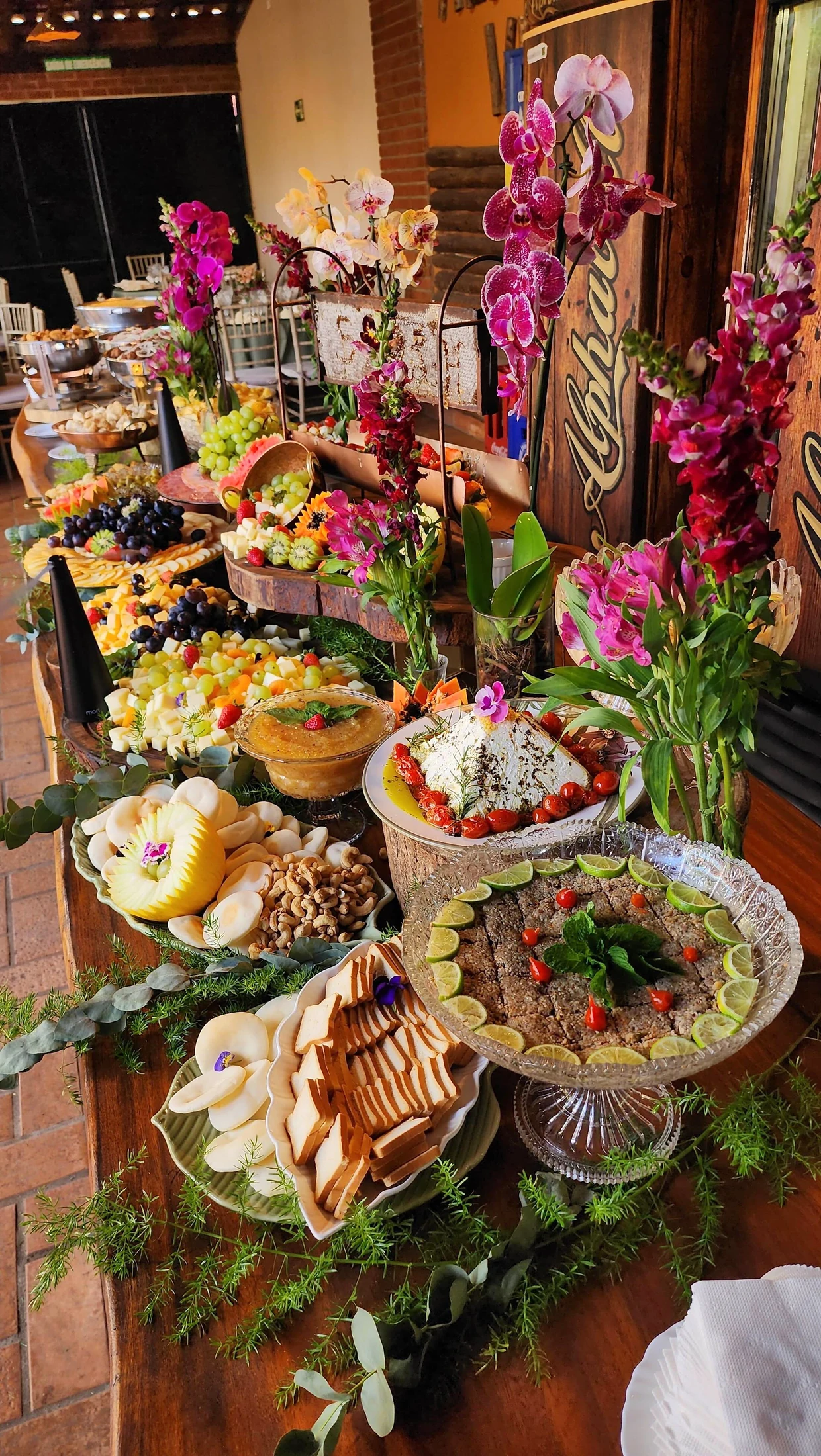 Mesa longa de frios com orquídeas e travessas de cristal montada para evento de empresa.