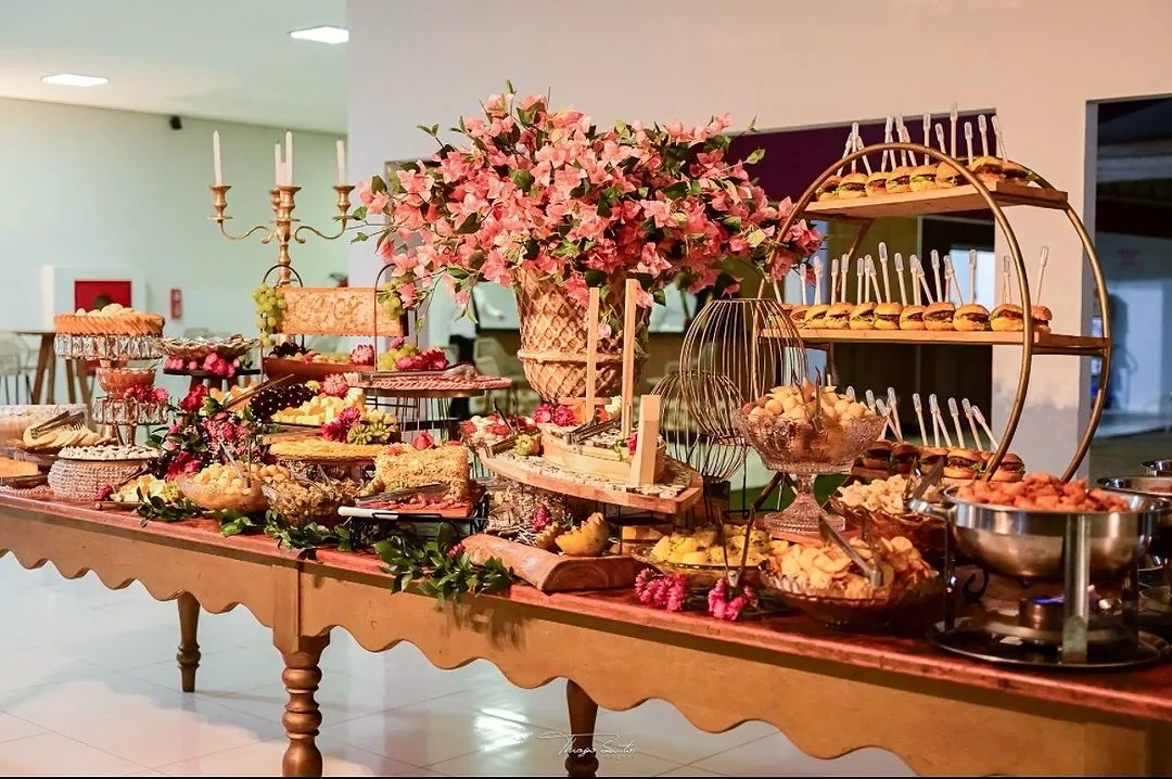 Mesa de buffet montada sob luz natural para recepção de convidados.